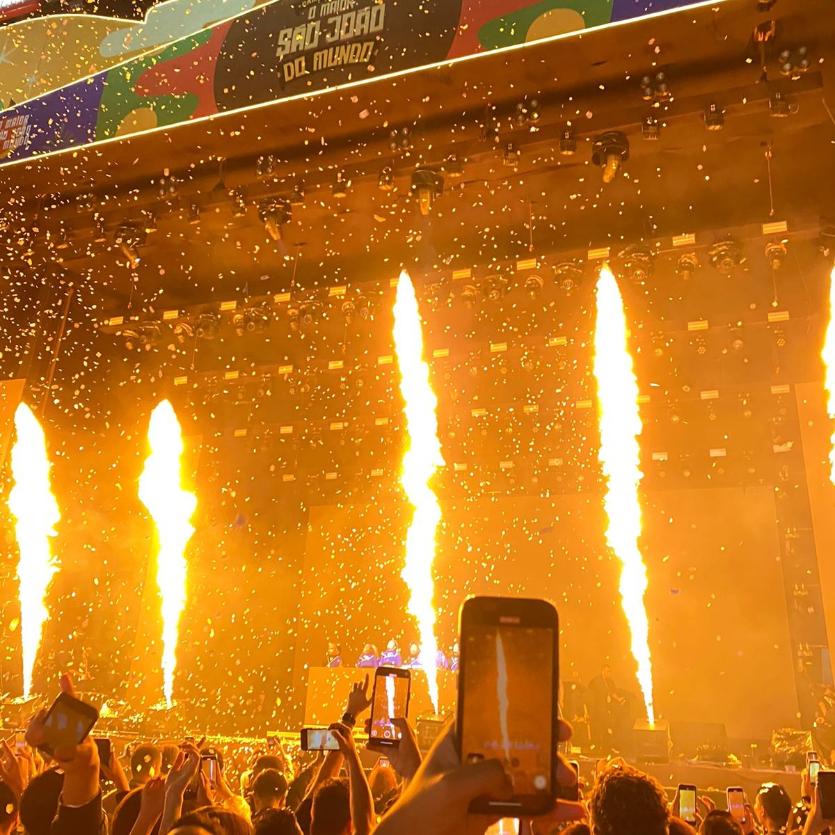 Muitas pessoas com celulares gravando um show. Em primeiro plano, uma mão segura um celular na vertical, gravando o palco. No palco, labaredas de fogo são lançadas verticalmente, enquanto confetes dourados brilham no ar. No topo do palco, um banner escrito 'O MAIOR SÃO JOÃO DO MUNDO' é visível.